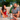 Close-up front view of a teal DankStop 7" Mini Bent Neck Bong held in hand against a blurred outdoor background with two people.