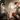 Two men seated on red chairs inhale from a tabletop Cipher NOVA Water Hookah Kit, shot from a frontal low angle showing the glass globe, twin flexible hoses, and vapor cloud.