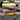Close-up of a yellow Cloud 8 Silicone Pizza Slice Hand Pipe shaped like a pepperoni pizza slice resting on a picnic table beside a red-checkered pizza box.